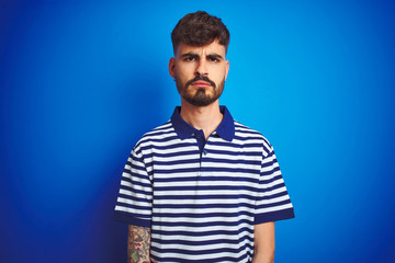 Fototapeta premium Young man with tattoo wearing striped polo standing over isolated blue background skeptic and nervous, frowning upset because of problem. Negative person.