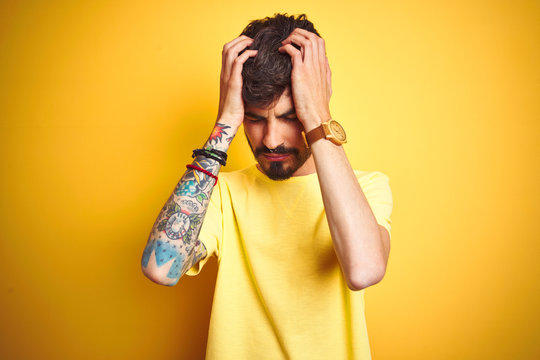 Young man with tattoo wearing t-shirt standing over isolated yellow background suffering from headache desperate and stressed because pain and migraine. Hands on head.