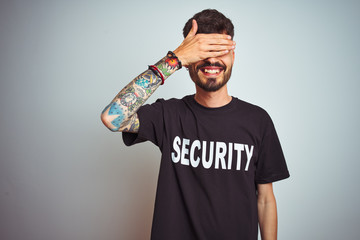 Young safeguard man with tattoo wering security uniform over isolated white background smiling and laughing with hand on face covering eyes for surprise. Blind concept.