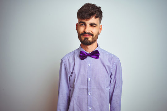 Young man with tattoo wearing purple shirt and bow tie over isolated white background winking looking at the camera with sexy expression, cheerful and happy face.