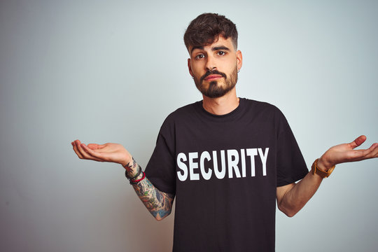 Young safeguard man with tattoo wering security uniform over isolated white background clueless and confused expression with arms and hands raised. Doubt concept.