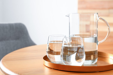 Tray with jug and glasses of water on wooden table in room, space for text. Refreshing drink