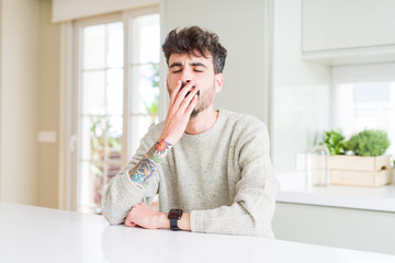 Fototapeta na wymiar Young man wearing casual sweater sitting on white table bored yawning tired covering mouth with hand. Restless and sleepiness.