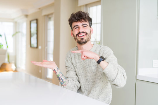 Young man wearing casual sweater sitting on white table amazed and smiling to the camera while presenting with hand and pointing with finger.