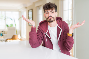 Young man wearing casual sweatshirt sitting on white table clueless and confused expression with...