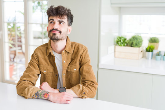 Young man wearing casual jacket sitting on white table smiling looking side and staring away thinking.