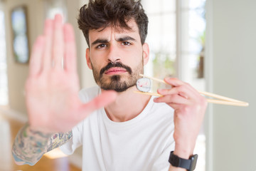 Young man eating asian sushi using chopsticks with open hand doing stop sign with serious and...