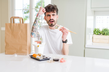 Young man eating asian sushi from home delivery annoyed and frustrated shouting with anger, crazy and yelling with raised hand, anger concept