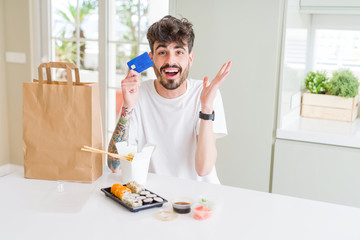 Young man eating asian sushi from home delivery using credit card as payment very happy and excited, winner expression celebrating victory screaming with big smile and raised hands