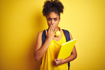 Afro woman using backpack and headphones holding notebook over isolated yellow background cover mouth with hand shocked with shame for mistake, expression of fear, scared in silence, secret concept