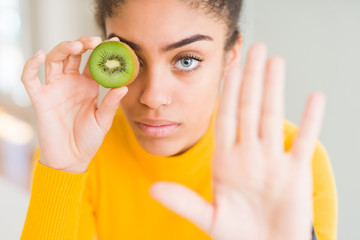 Young african american girl eating green kiwi with open hand doing stop sign with serious and confident expression, defense gesture