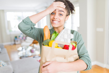 Young african american girl holding paper bag of groceries from supermarket stressed with hand on head, shocked with shame and surprise face, angry and frustrated. Fear and upset for mistake.