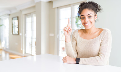 Beautiful young african american woman with afro hair sitting on table at home showing and pointing up with finger number one while smiling confident and happy.