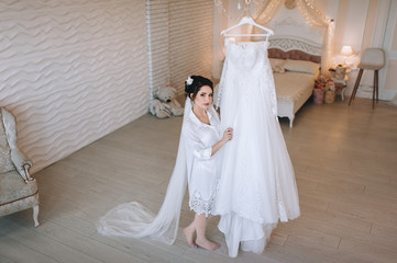 Plakat A beautiful bride in a bathrobe, lingerie and a long veil stands in the studio, and holds a white dress on a hanger. Wedding portrait of a cute girl in the studio. Photography and concept.