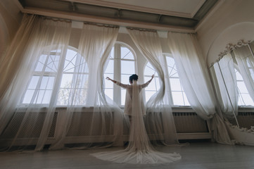 A beautiful bride in a bathrobe and lingerie stands near the window and holds on to the curtains, pushing them apart. Wedding portrait of a cute girl with a long veil. Photography and concept.