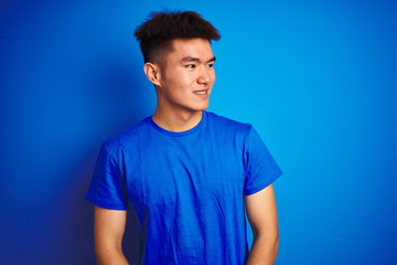 Young asian chinese man wearing t-shirt standing over isolated blue background looking away to side with smile on face, natural expression. Laughing confident.