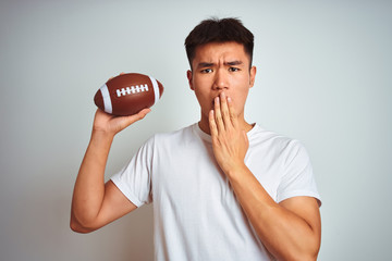 Young asian chinese sportsman holding rugby ball standing over isolated white background cover mouth with hand shocked with shame for mistake, expression of fear, scared in silence, secret concept