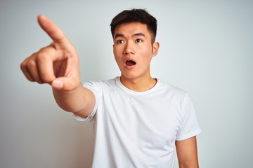 Young asian chinese man wearing t-shirt standing over isolated white background Pointing with finger surprised ahead, open mouth amazed expression, something on the front