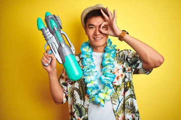 Asian chinese man wearing floral hawaian lei and water gun over isolated yellow background with happy face smiling doing ok sign with hand on eye looking through fingers