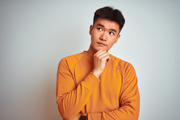 Young asian chinese man wearing orange sweater standing over isolated white background with hand on chin thinking about question, pensive expression. Smiling with thoughtful face. Doubt concept.