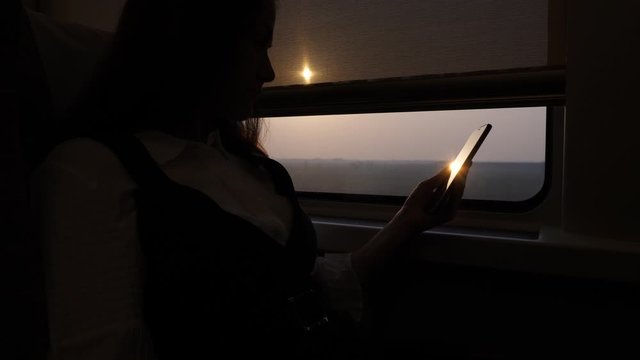 Woman sit with phone in hand against train window, shaded inside, evening sun shine outside, flickering reflection at glass of smartphone screen. Business lady using modern high speed train to travel