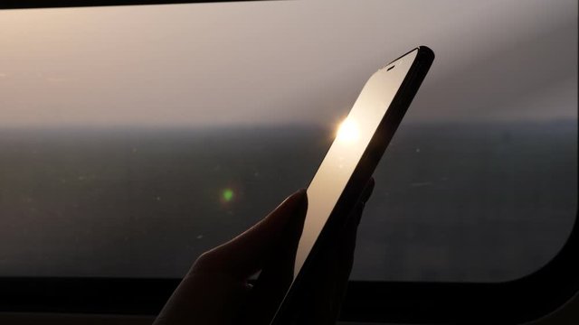 Bright sun light reflection on glass of phone screen, high contrast close up shot. Passenger hold smartphone against window, high speed train running at countryside, blurred background