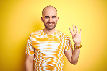 Young bald man with beard wearing casual striped t-shirt over yellow isolated background showing and pointing up with fingers number four while smiling confident and happy.