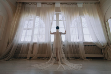 A beautiful bride in a bathrobe and lingerie stands near the window and holds on to the curtains, pushing them apart. Wedding portrait of a cute girl with a long veil. Photography and concept.