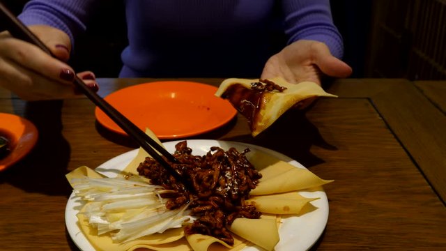 European woman try to eat Chinese food, put sauteed pork strips on sheet of tofu using chopsticks, then add white onion. Popular Beijing style meal, Jing Jiang Rou Si, served at restaurant