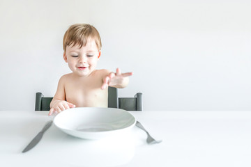 Cute blond boy sitting at the table with an empty white plate, fork and knife.