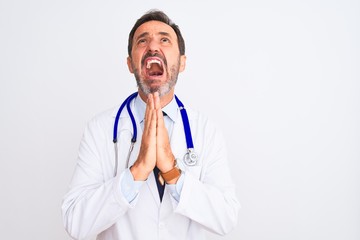 Fototapeta premium Middle age doctor man wearing coat and stethoscope standing over isolated white background begging and praying with hands together with hope expression on face very emotional and worried