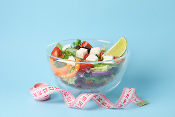 Glass bowl with fresh salad and measuring tape on blue background, space for text