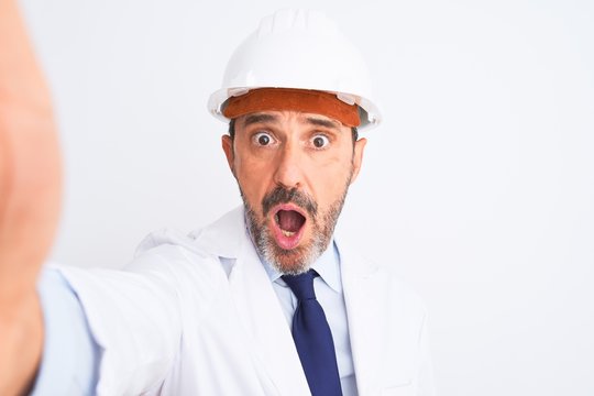 Middle age engineer man wearing helmet make selfie over isolated white background scared in shock with a surprise face, afraid and excited with fear expression