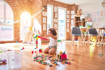 Beautiful toddler wearing glasses and unicorn diadem playing with tractor, building blocks toy and tennis racket at kindergarten