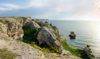 Fototapeta na wymiar Sea coast with steep textured limestone rocks and shipwrecked ship