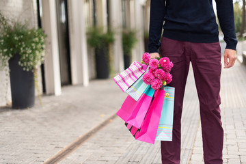Stylish man holding flowers and pink, blue shopping bags near restaurant. Woman's day. Valentine's day. Ready for birthday party or romantic date. Copy space. Bunch of paper gift bags