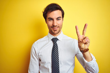 Young handsome businessman wearing elegant shirt and tie over isolated yellow background showing and pointing up with fingers number two while smiling confident and happy.
