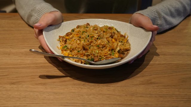 Woman hold medium plate with stir fried rice with meat and vegetables, typical meal at Chinese restaurant. She demonstrate freshly cooked food then take spoon and scoop rice