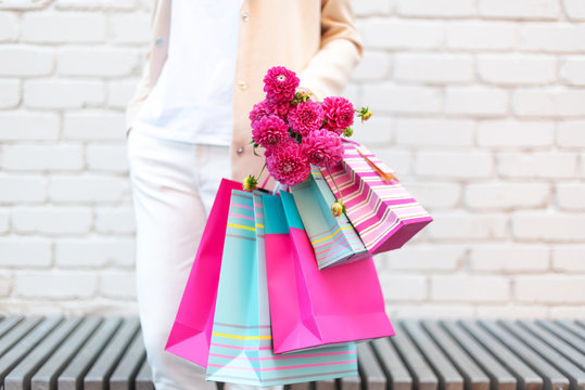 Pink flowers in female hands and shopping bags on brick background. Birthday, Mother's, Women's, Wedding Day concept. Bunch of paper gift bags