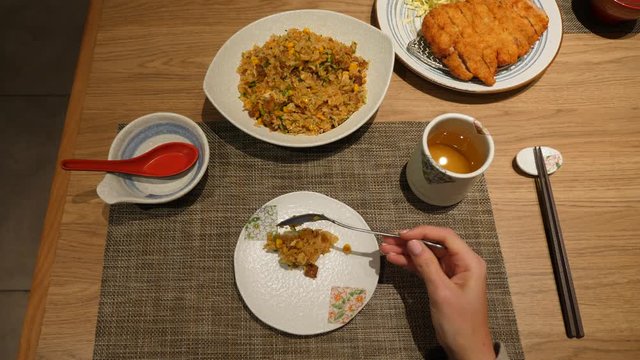 Woman take several spoons of rice from shared plate, top view of table at Chinese restaurant. She put stir fried rice on personal dish, cup of tea and breaded sliced meat meal stay near