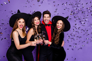 Photo of three witch ladies and wizard guy at helloween event drink golden wine glitter in air wear black dresses caps suit and long coat isolated purple color background