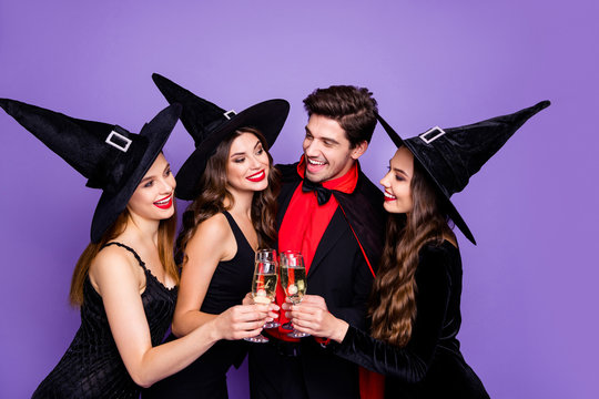 Photo of three excited witch ladies and warlock guy chilling at halloween party drink golden wine wear black dresses hats and vampire coat isolated purple color background