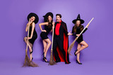 Full body photo of three witch ladies and wizard guy rock band holding brooms singing songs chilling wear black dresses caps suit and long coat isolated purple color background