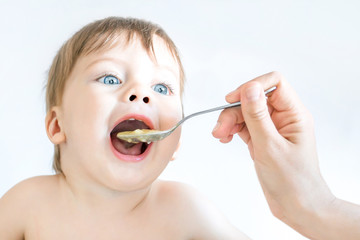 Cute blue-eyed baby is eating with a spoon, with mom's help.