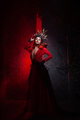 Fototapeta na wymiar beautiful fashion witch woman with horns in the form of tree roots in a long luxurious dress on the background of a huge gate with red smoke. Halloween concept
