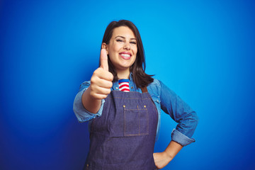 Young beautiful business woman wearing store uniform apron over blue isolated background doing happy thumbs up gesture with hand. Approving expression looking at the camera with showing success.