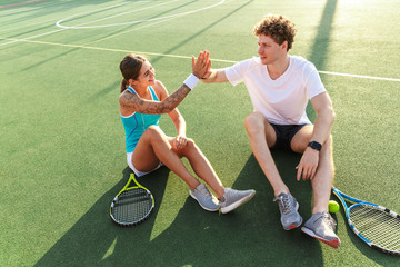 Image of beautiful man and woman sitting on tennis court outdoors