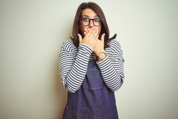 Young beautiful business woman wearing store uniform apron over isolated background shocked covering mouth with hands for mistake. Secret concept.