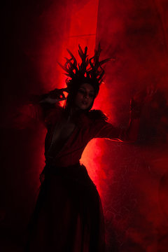beautiful fashion witch woman with horns in the form of tree roots in a long luxurious dress on the background of a huge gate with red smoke. Halloween concept