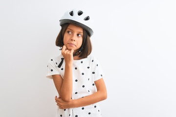 Beautiful child girl wearing security bike helmet standing over isolated white background with hand on chin thinking about question, pensive expression. Smiling with thoughtful face. Doubt concept.